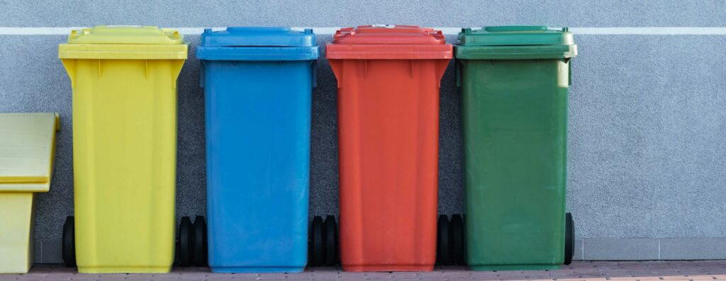 four assorted-color trash bins beside gray wall